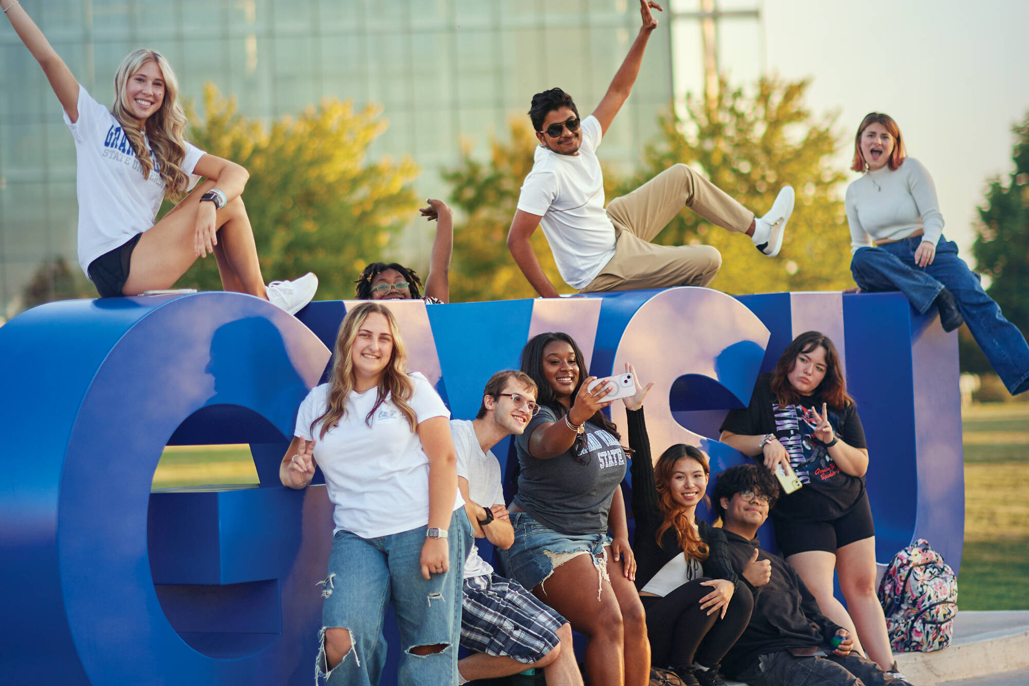 A group of students sitting or leaning on the big GVSU letters in the lawn in front of the library, smiling for the camera